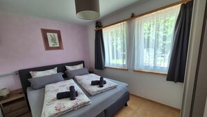 Ein Schlafzimmer mit Bett, Nachttisch, Lampe, einem Fenster mit Vorhängen und einem Bild an der Wand.