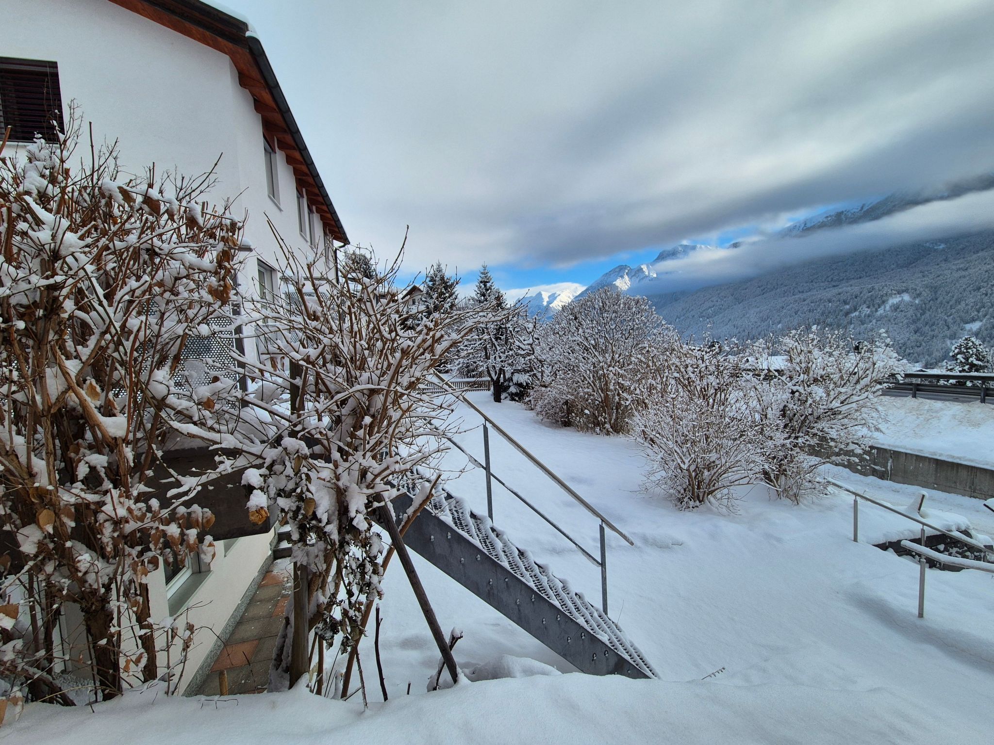 A snow-covered outdoor area with a staircase leading to a building, surrounded by snow-laden trees and a mountainous backdrop.