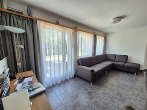 A living room with a sectional sofa, a wooden table with magazines, and a window with sheer curtains.