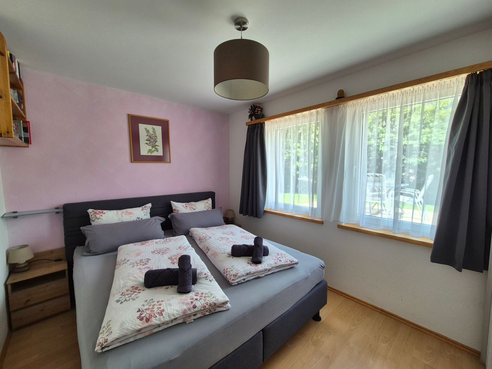 A bedroom with a bed, a bedside table, a lamp, a window with curtains, and a wall-mounted picture.