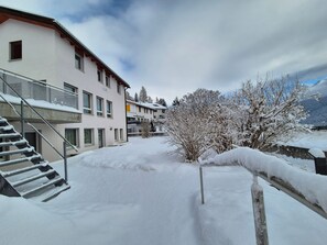Une cour enneigée avec un escalier menant à un bâtiment, le tout entouré d’arbres et de montagnes.
