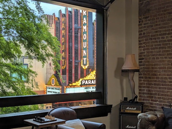 A room with a view of a theater marquee, a table with chess pieces, a leather couch, a lamp, and a Marshall amplifier.