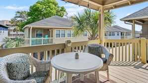 Ein überdachter Außenbereich mit rundem Tisch und Korbstühlen, einer Holzterrasse und Blick auf ein Haus mit Balkon.