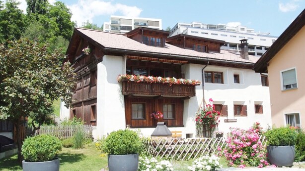 Un edificio in stile chalet tradizionale con balcone in legno, piante in vaso e vialetto sterrato.