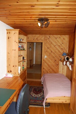 Ett litet rum med en säng, ett skrivbord och en stol. Det finns en garderob i trä med hyllor, en taklampa och en dörr som leder till ett annat rum.