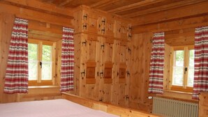 Quarto de cabana de madeira com cama, duas janelas com cortinas e um radiador.