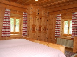 A wooden cabin bedroom with a bed, two windows with curtains, and a radiator.
