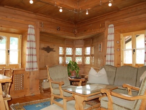 A wooden cabin interior with a dining table, chairs, a sofa, and a potted plant.