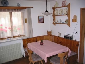 A dining area with a table and chairs, a radiator, a window with curtains, a shelf with dishes, and a radio.