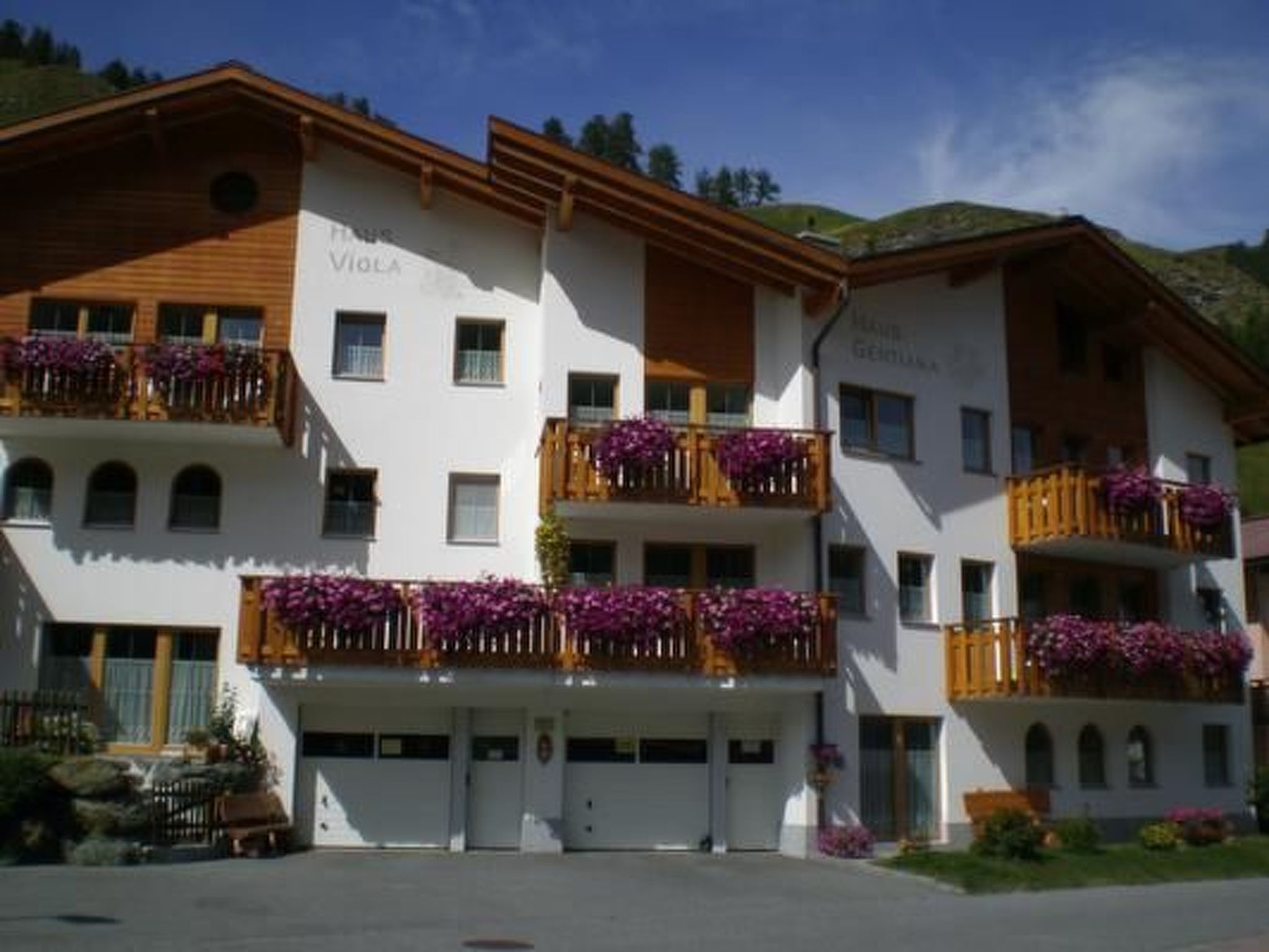 Et hotel med flere etager, hvor der er altaner pyntet med lilla blomster, en hvid facade og et bjergrigt landskab i baggrunden.