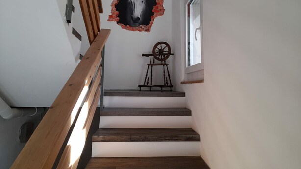 Uma escadaria com degraus de madeira e corrimão, que leva a um quarto com janela e um objeto fixado na parede.