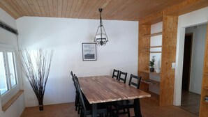 Uma área de refeições com mesa e cadeiras de madeira, luminária pendente, quadro na parede e planta em um vaso sobre uma prateleira.