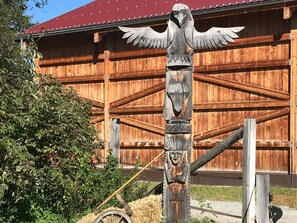 A totem pole with wings and a carved face, standing in front of a wooden building with a red roof.