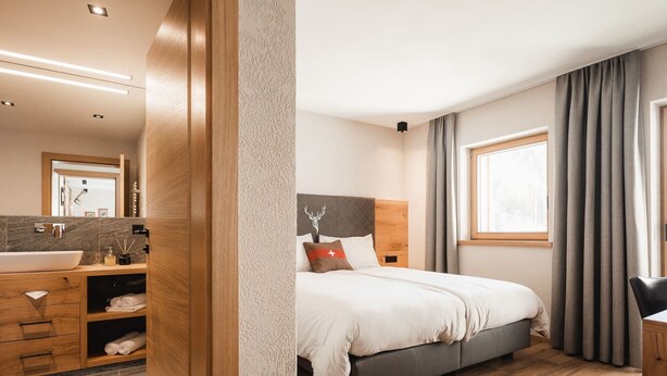 A modern hotel room with a wooden floor, a large bed with a headboard, a bathroom with a sink and mirror, and a window with curtains. - Avero Lodge by MEIN SAMNAUN – MOUNTAIN HOMES, (Samnaun-Compatsch) by Interhome (Samnaun)