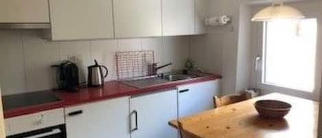 Kompakti keittiö, jossa on valkoiset kaapit, punainen työtaso ja puinen ruokapöytä.