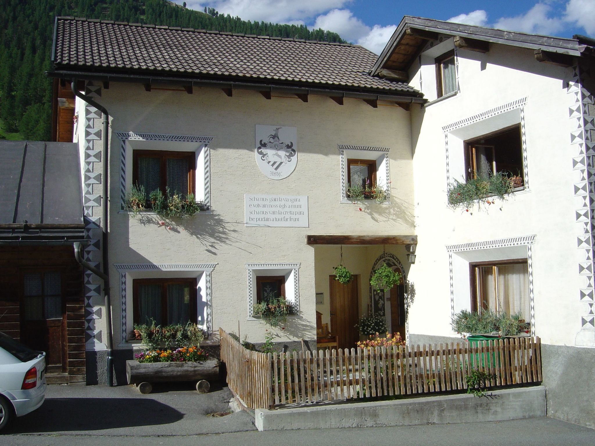 A two-story building with a tiled roof, a coat of arms, and a sign with text. There are windows with plants, a wooden fence, and a car parked in front.