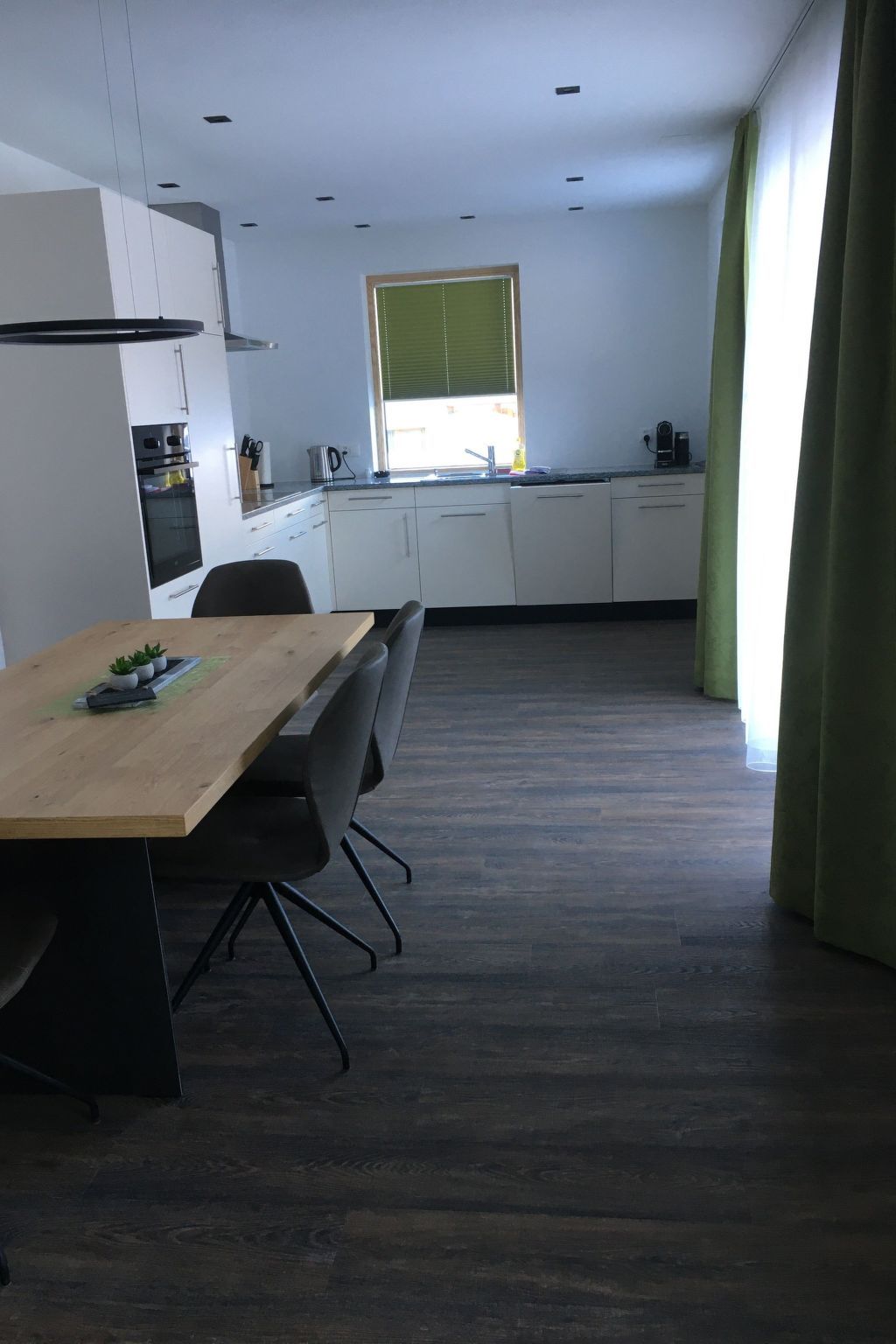 A modern kitchen with white cabinets, a wooden dining table, and a window with blinds.
