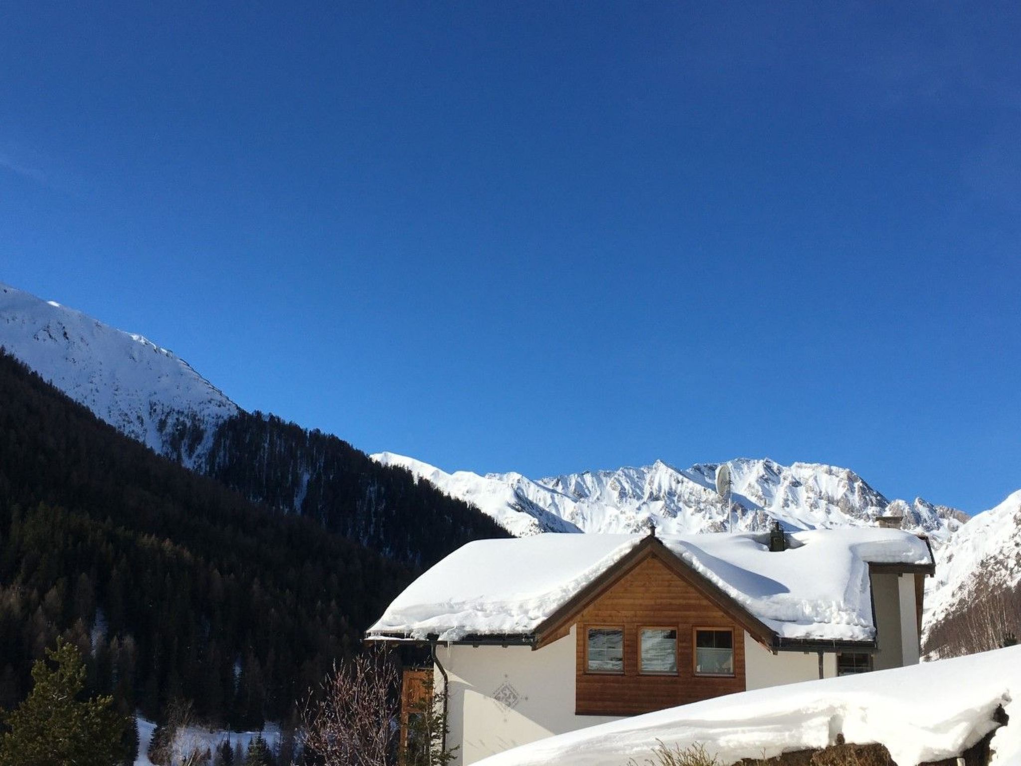 A wooden chalet with a sloped roof, surrounded by snow-covered mountains and a clear blue sky.