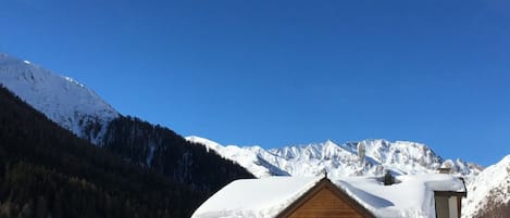 A wooden chalet with a sloped roof, surrounded by snow-covered mountains and a clear blue sky.