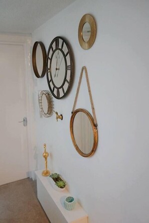 A hallway with a white door, a white bench, and a wall adorned with various mirrors and a small golden lamp. - One Bed Apartment in Clifton (Bristol)