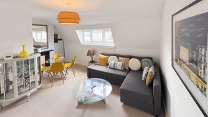 Ein Wohnzimmer mit einer Couch, einem Glastisch, einem Essbereich mit Stühlen, einem Schrank und einem großen Fenster.