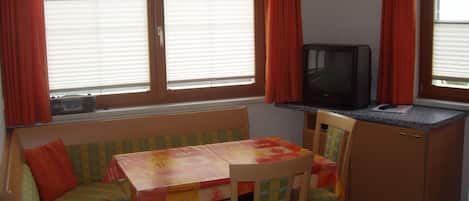 Una cocina compacta con estufa integrada, una pequeña mesa de comedor con mantel colorido y un televisor sobre un mueble.
