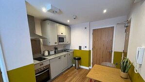 A compact kitchen with a microwave, stove, and sink, a dining table with chairs, and a small trash bin. - Prince Street Studio 7 (Bristol)