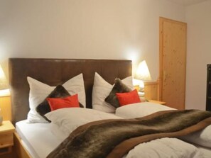 Ein Bett mit dunklem Kopfteil, weißer Bettwäsche und mehreren Kissen. Auf den hölzernen Nachttischen stehen je eine Nachttischlampe.