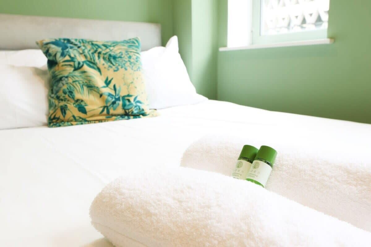 A bed with white bedding, a blue and green pillow, and two bottles on the bed.