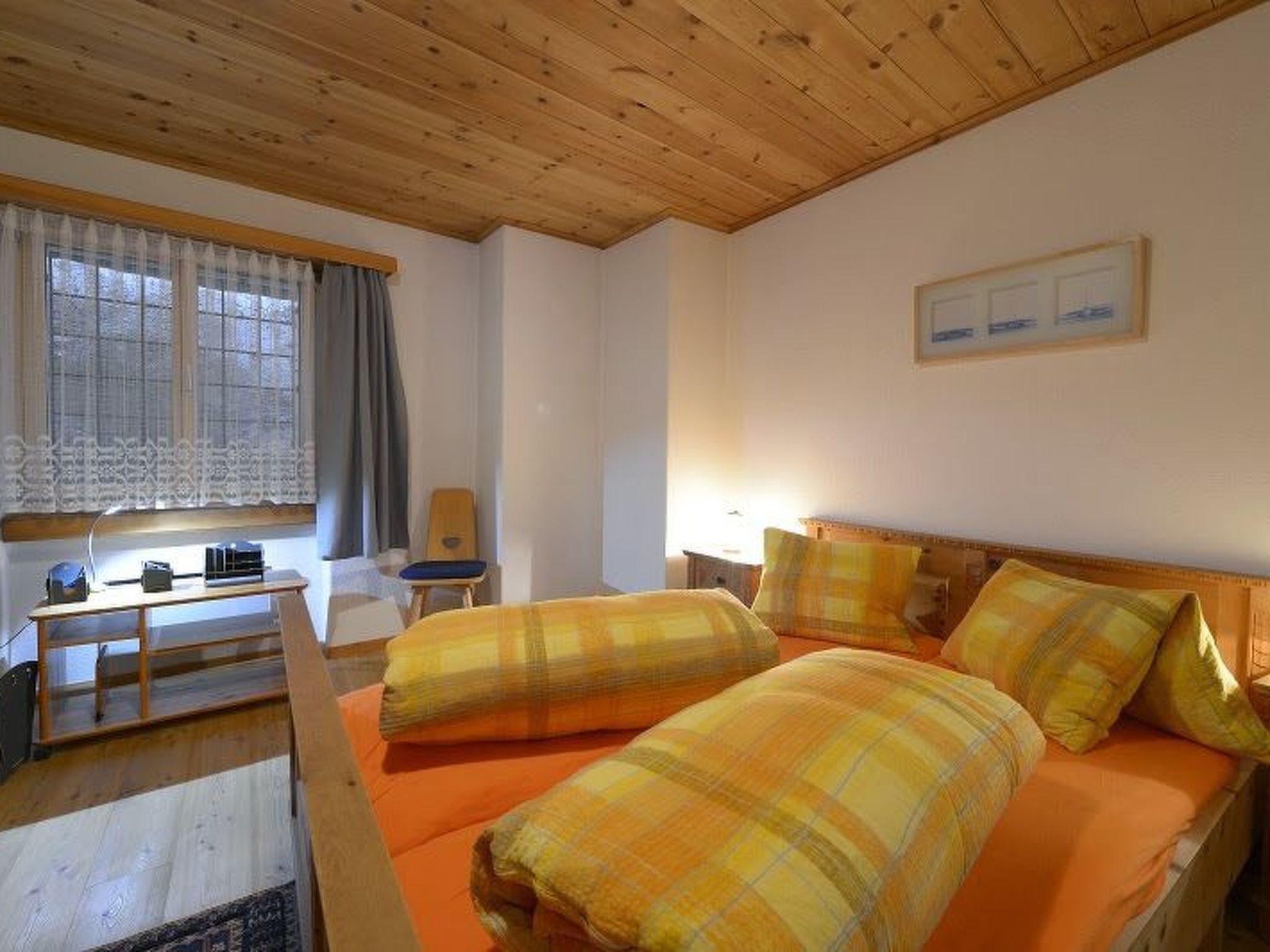 A room with a wooden ceiling, a bed with orange bedding, a desk with a chair, and a window with curtains.