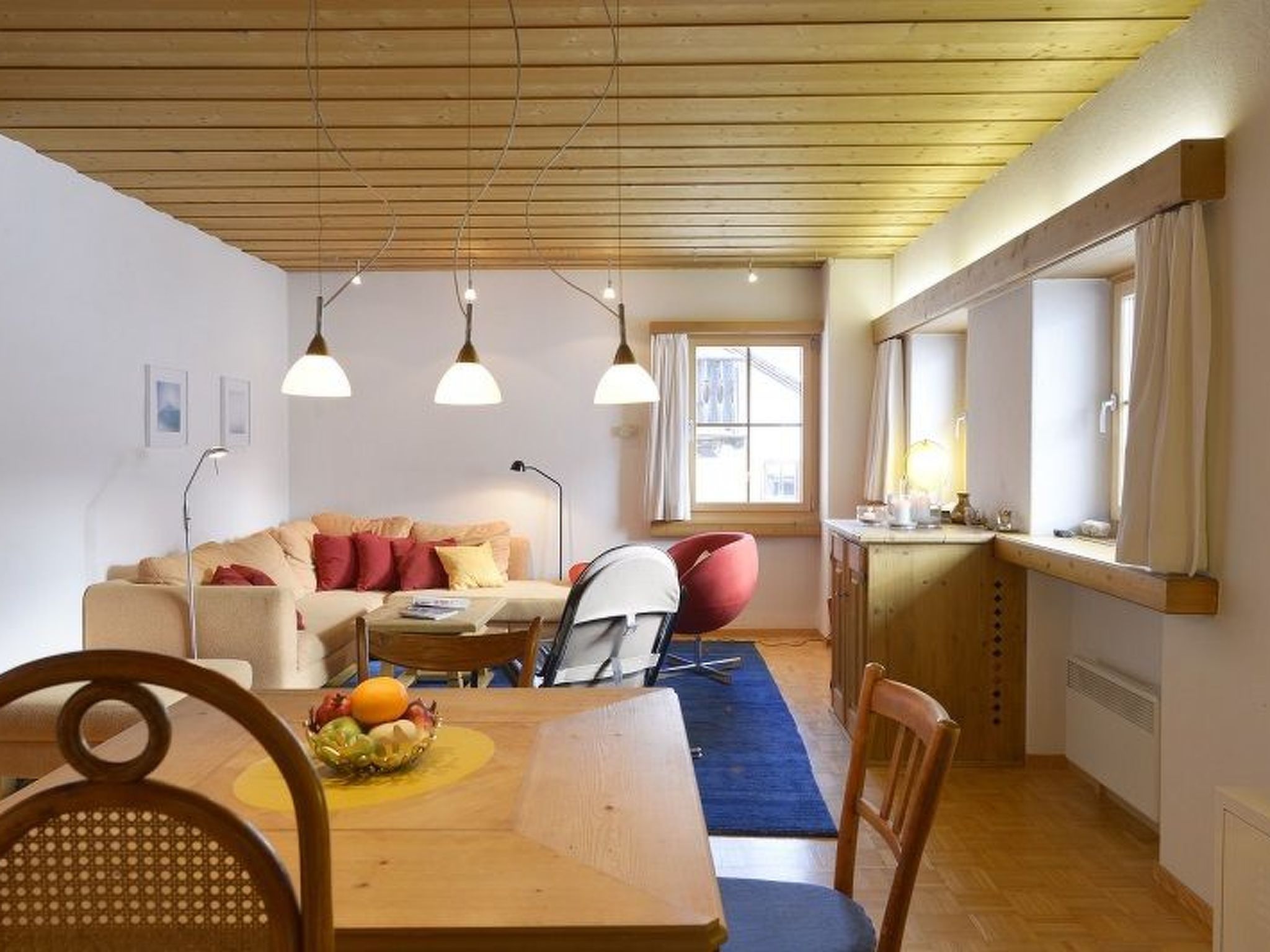 A cozy living room with a wooden ceiling, a dining table with chairs, a sofa, and a red chair.