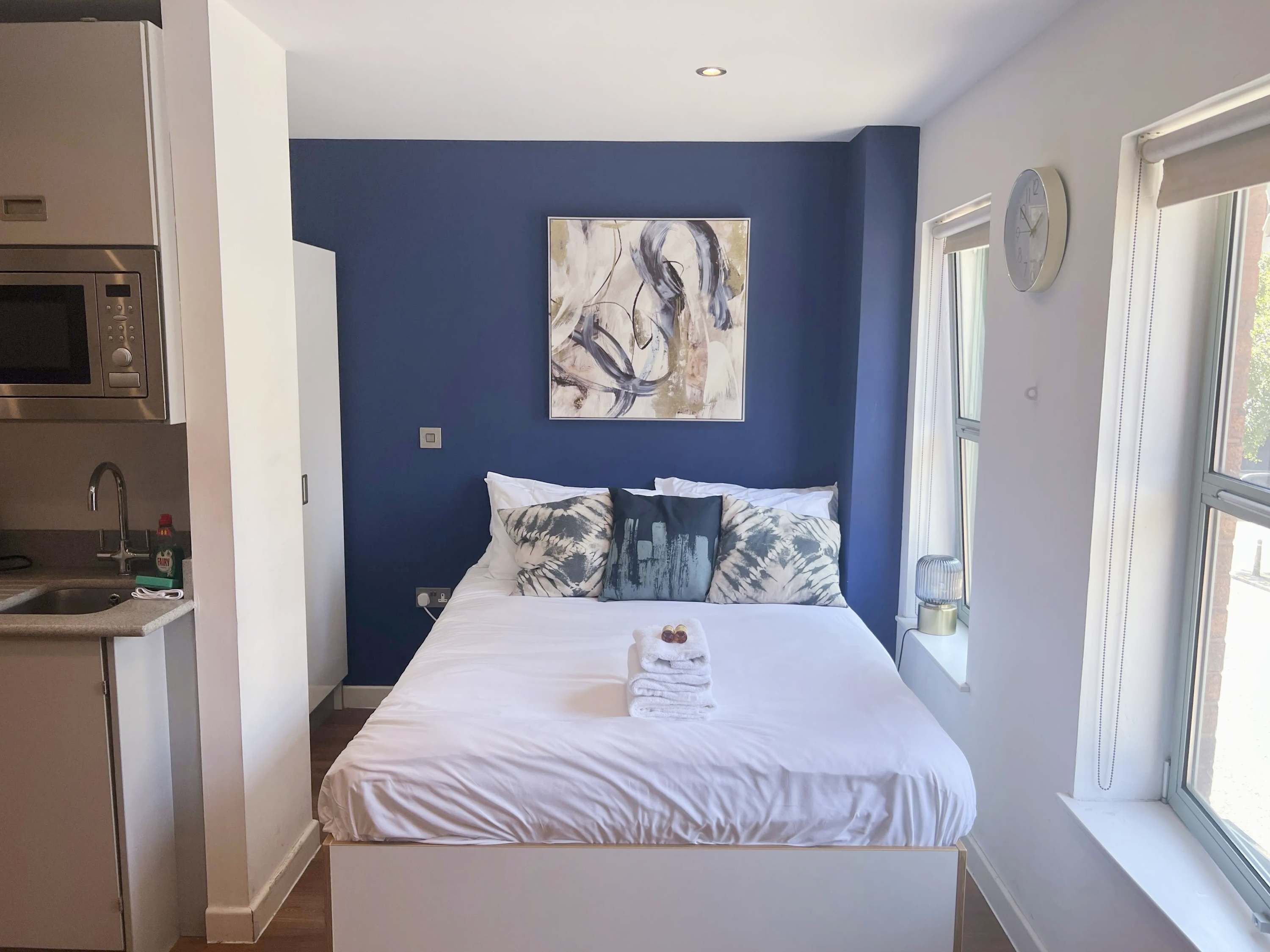A bedroom with a bed, pillows, a painting, a clock, and a window.