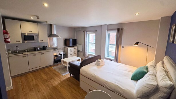 Una habitación de hotel pequeña con kitchenette, cama, mesita de noche y ventana.