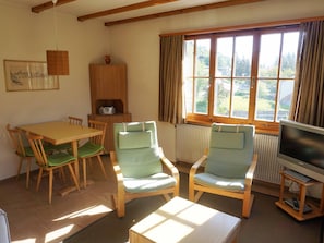 A furnished living room with a dining table, two armchairs, a television, and a window with curtains. - Ferienhaus Chasa Allegra, (Scuol) by Interhome (Scuol)