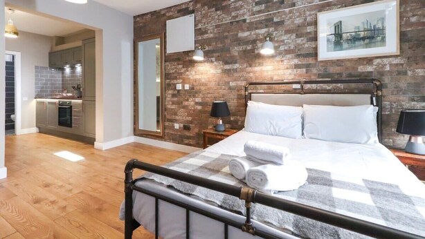 A modern bedroom with a metal bed frame, a large window, and a brick wall. - Old Market Studios-No1 (City of Bristol)