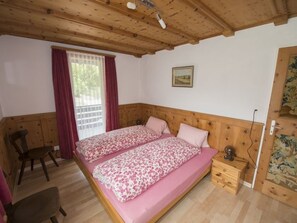 Ein Doppelbett mit floralem Bettzeug, ein hölzernes Kopfende, ein Fenster mit Vorhängen, ein Stuhl und ein kleiner Nachttisch.