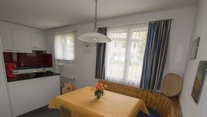 A compact kitchen with white cabinets and a red appliance, a dining area with a yellow table and blue bench, and a window with curtains. - STEINMANN, (Scuol) by Interhome (Scuol)