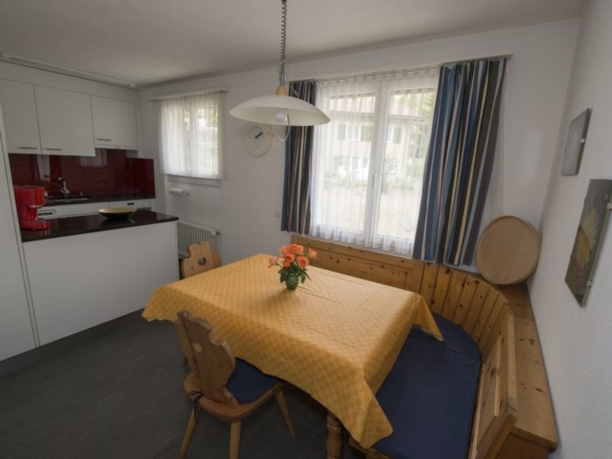 A compact room with a dining table, a wooden bench, a small kitchen area, and a window with curtains.
