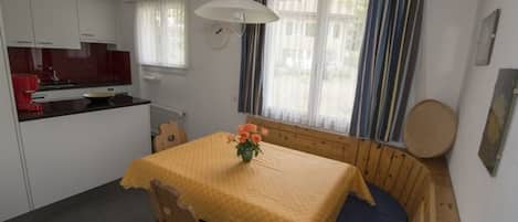 A compact room with a dining table, a wooden bench, a small kitchen area, and a window with curtains.