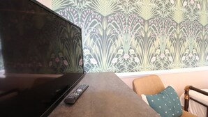 Een televisie op een tafel met een afstandsbediening, behang met een patroon en een stoel met een kussen.