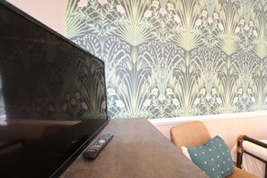 A television on a table with a remote control, a patterned wallpaper, and a chair with a pillow. - Alma Studios - Apartment 4 - Hopewell (Bristol)