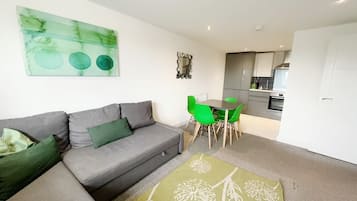 A modern living room with a grey sofa, green cushions, a green dining table with chairs, and a kitchen in the background.