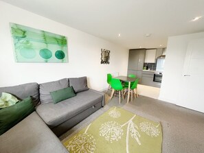 Una sala moderna con un sofá gris, cojines verdes, una mesa de comedor verde con sillas y una cocina al fondo.