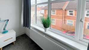 Ein Zimmer mit großem Fenster, einem Heizkörper, einem weißen Nachttisch mit Lampe und einer Topfpflanze.