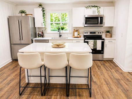 A modern kitchen with a central island, three bar stools, stainless steel appliances, and a window with greenery.. Villa Playera a 20 minutos del uptown de Charlotte frente al Lago