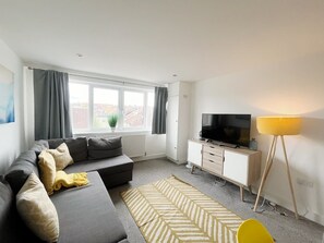 A modern living room with a sofa, television, cabinet, lamp, and a view of the city through large windows.