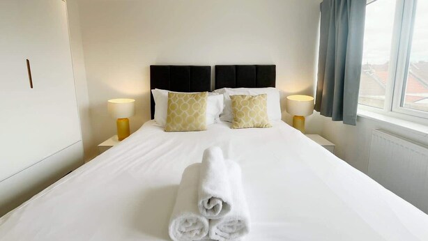 A hotel room with a large bed, two bedside lamps, a rolled towel on the bed, and a window with curtains.