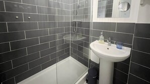 A bathroom with dark tiled walls, a white bathtub, and a sink with a mirror above it. - Anstey Heights - Apartment 3 - Hopewell (Easton)