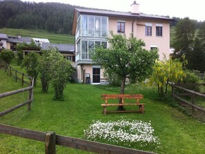 A two-story building with a balcony, surrounded by a wooden fence and a grassy area with a bench and white flowers. - Iten, (Ftan) by Interhome (Ftan)
