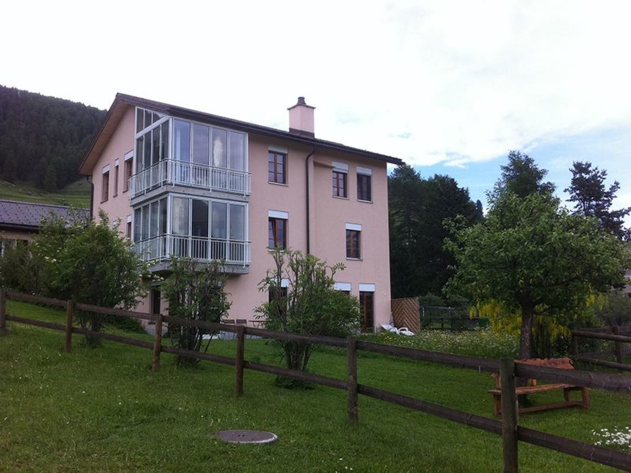 A multi-story building with a balcony, surrounded by a wooden fence and greenery.